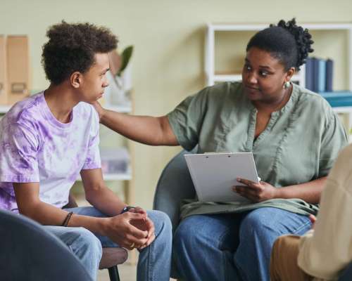 African young psychologist talking to teenage boy during therapy session at classroom