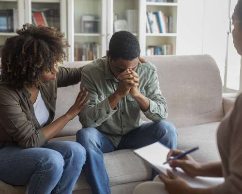 Loving attentive young african american woman hugging comforting her depressed spouse or boyfriend suffering from anxiety, black couple having family therapy with woman psychologist, clinic interior